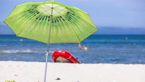 Ein Kiwi-Sonnenschirm steht am Strand, im Hintergrund sind Badegäste und ein Schlauchboot zu sehen.