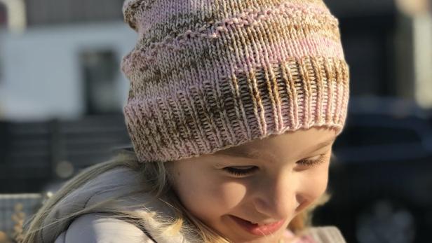 Ein lächelndes Mädchen trägt eine rosa-braun gestreifte Mütze und eine graue Jacke.