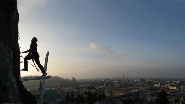 Eine Kletterin mit Helm an einer Felswand mit Blick auf die Stadt Salzburg.