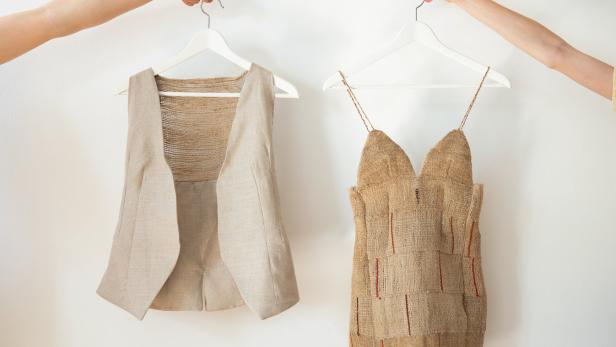 Zwei Hände halten eine Weste und ein Kleid aus grobem Stoff an Kleiderbügeln vor einer weißen Wand.