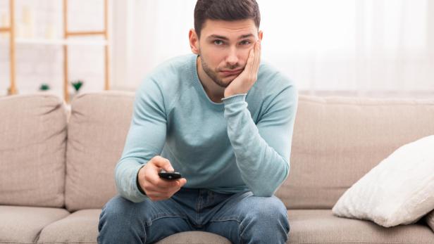 Ein Mann sitzt gelangweilt auf einem Sofa, hält eine Fernbedienung und stützt den Kopf auf die Hand.