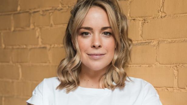 Eine Frau mit blonden Haaren und einem weißen T-Shirt posiert vor einer gelben Backsteinmauer.
