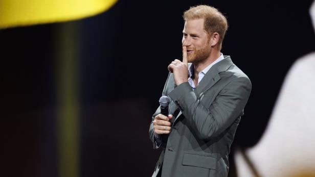 Prinz Harry hält ein Mikrofon und deutet mit dem Finger zum Schweigen.