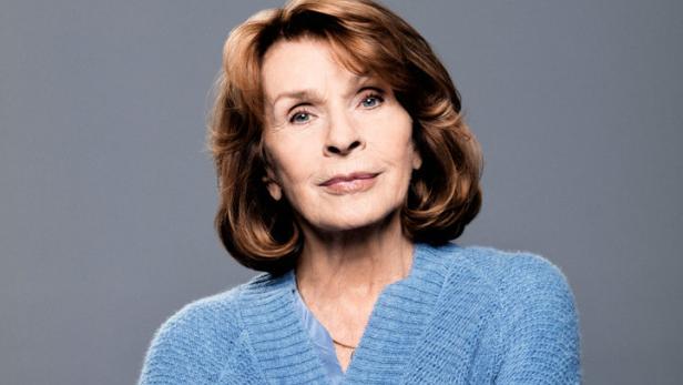 Eine ältere Frau mit verschränkten Armen trägt eine blaue Strickjacke.