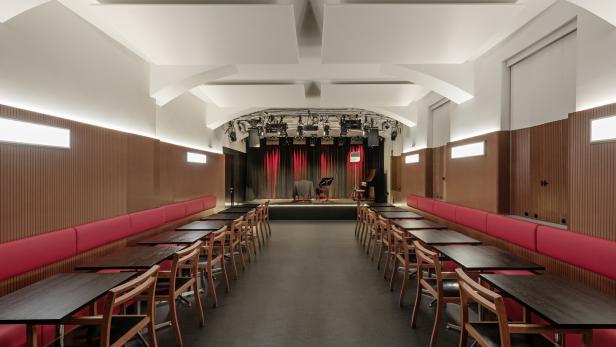 Ein leerer Veranstaltungsort mit Tischen und Stühlen vor einer Bühne mit rotem Vorhang.