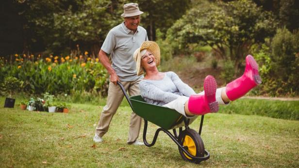 Älterer Mann schiebt glückliche Frau in der Schubkarre. Sie streckt die Füße in pinken Gummistiefeln lachend in die Luft.