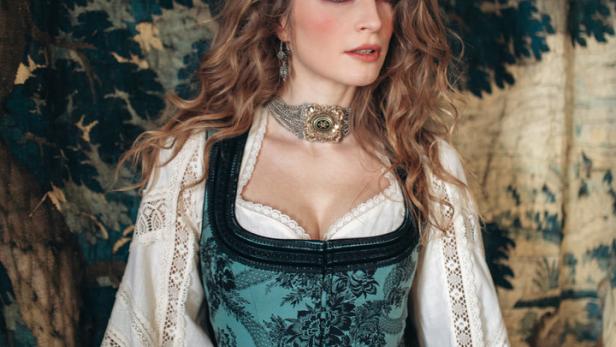 Eine Frau in Tracht vor einem Wandteppich mit Baumdarstellung.