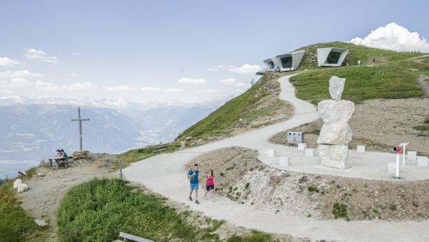 Ein Paar wandert auf einem Bergpfad mit Blick auf ein modernes Gebäude und eine Berglandschaft.
