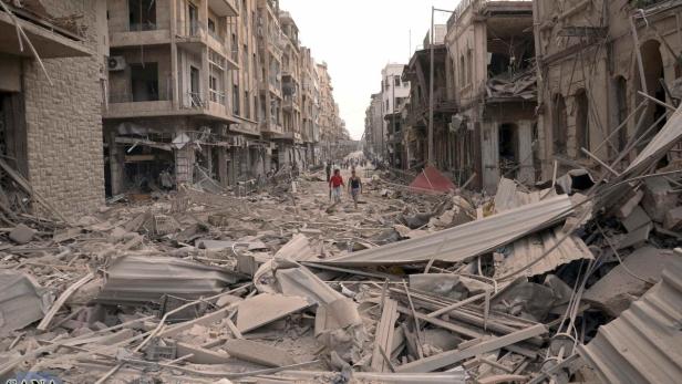 Zwei Personen gehen durch eine von Trümmern übersäte Straße mit zerstörten Gebäuden.