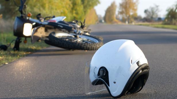 Ein weißer Motorradhelm liegt auf einer Straße, im Hintergrund liegt ein umgefallenes Motorrad.