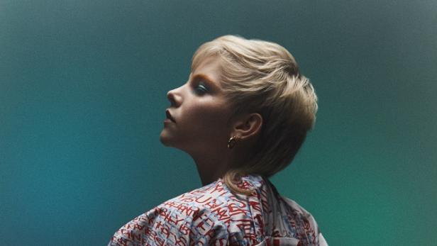 Eine Frau mit kurzer, blonder Herbstfrisur und einem Anzug mit Schriftmuster blickt nach oben.