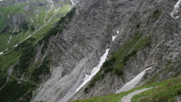Ein Bergpfad schlängelt sich an einem steilen, felsigen Hang entlang, mit Schneeresten dazwischen.