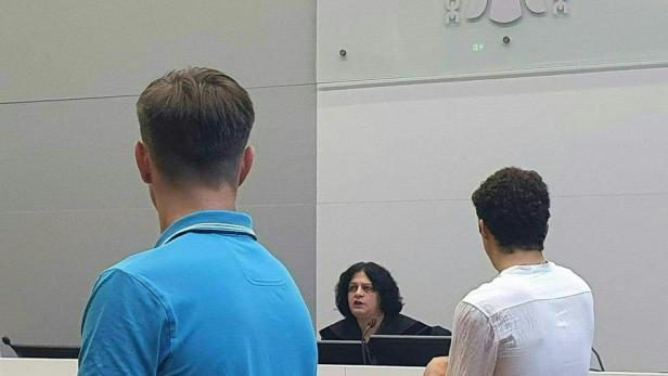 Zwei Männer stehen vor einer Richterin im Gerichtssaal, im Hintergrund das Justiz-Logo.