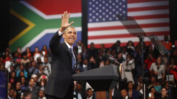 Barack Obama winkt von einem Rednerpult vor Flaggen Südafrikas und der USA.