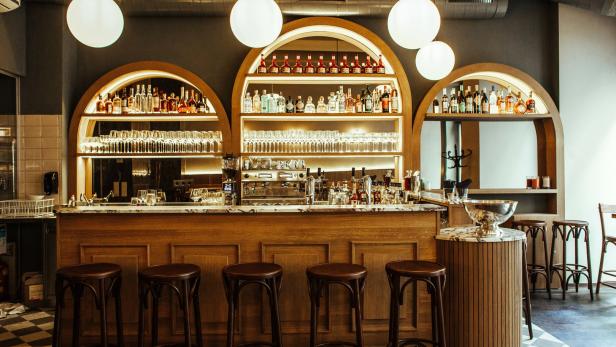 Eine Bar mit hölzernen Hockern und Regalen voller Flaschen und Gläser.