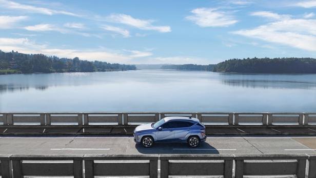 Ein blauer Hyundai Kona fährt über eine Brücke vor einer Seenlandschaft.