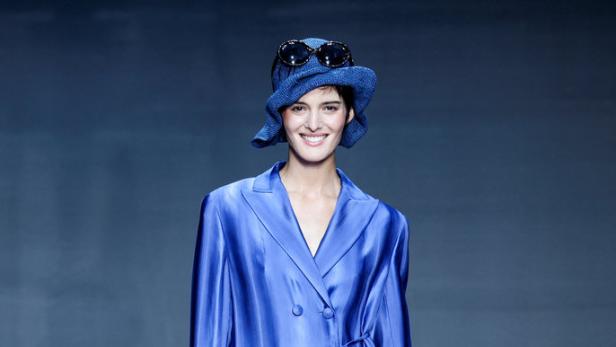 Eine Frau in einem blauen Blazer, einem Hut und einer transparenten Hose mit floralen Applikationen auf einem Laufsteg.