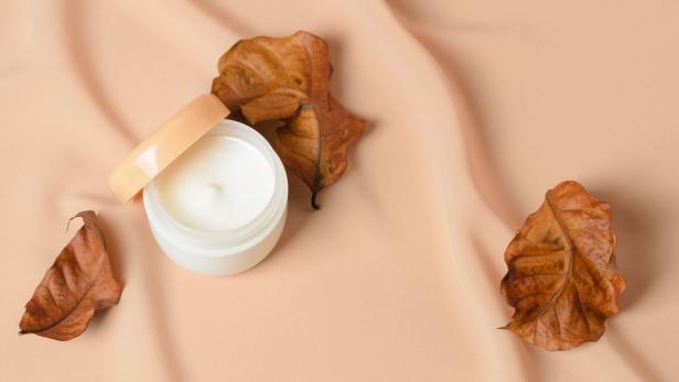 Eine geöffnete Dose mit Hautcreme liegt neben trockenen Blättern auf einem beigen Tuch.