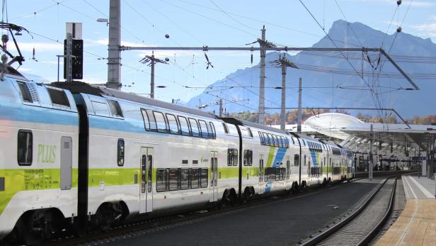 Ein doppelstöckiger Westbahn-Zug steht an einem Bahnhof vor einer Bergkulisse.
