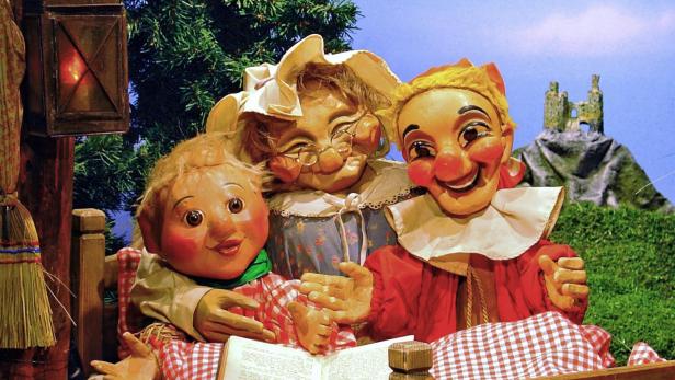 Drei Puppen sitzen in einem Bett und lesen ein Buch vor einer gemalten Landschaft.