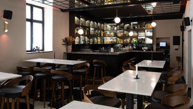 Innenansicht einer Bar mit Tischen, Stühlen und einer beleuchteten Bar im Hintergrund.