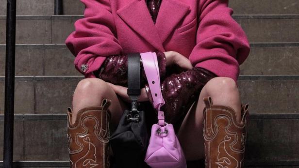 Eine Frau in Cowboystiefeln und pinkfarbenem Blazer sitzt auf einer Treppe und hält zwei Handtaschen.