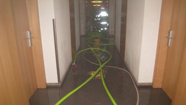 Ein überfluteter Flur mit Feuerwehrleuten im Hintergrund und Schläuchen auf dem Boden.