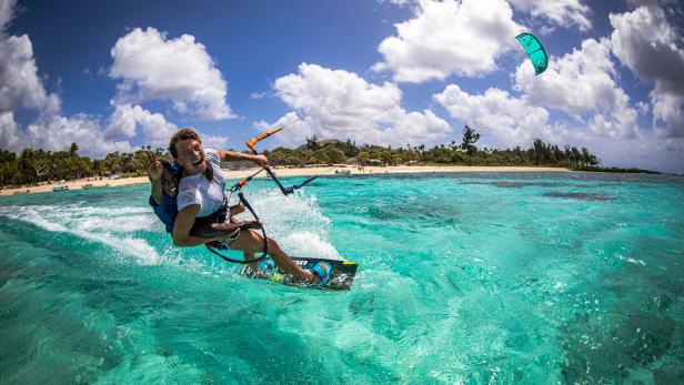 Zwei Personen beim Kitesurfen auf türkisfarbenem Wasser vor einer tropischen Küste.