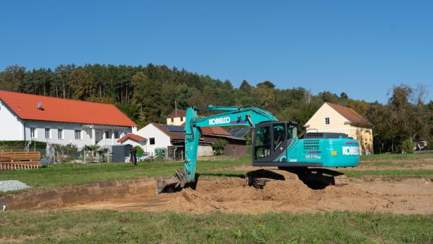 Ein türkisfarbener Bagger hebt Erde auf einer Baustelle vor Wohnhäusern und Bäumen aus.