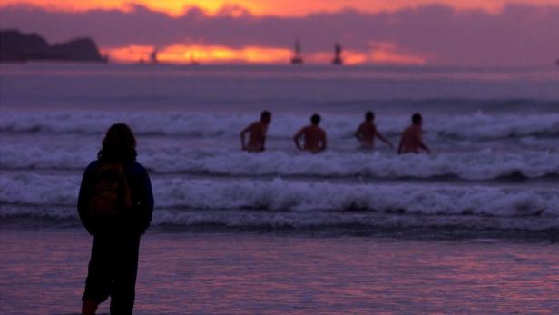 Eine Person mit Rucksack steht am Strand, während andere im Meer schwimmen.