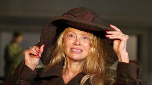 Pamela Anderson mit blonden Haaren und rotem Nagellack trägt einen braunen Hut und Mantel.