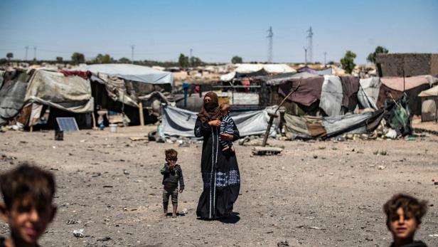 Eine verschleierte Frau trägt in einem Flüchtlingscamp ein Kleinkind, wird von einem anderen begleitet