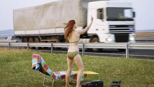 Eine Frau im Bikini steht an einer Autobahn und zeigt einem LKW den Stinkefinger.