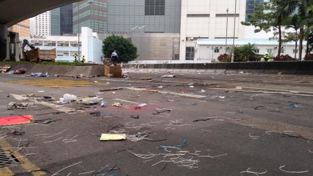 Eine Straße ist mit Müll und Trümmern übersät; im Hintergrund sind Aufräumarbeiten zu sehen.