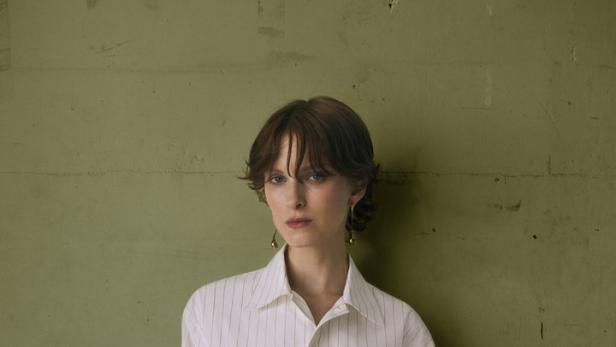 Eine Frau mit Ohrringen posiert in einem gestreiften Hemd und einem schwarzen Rock vor einer grünen Wand.