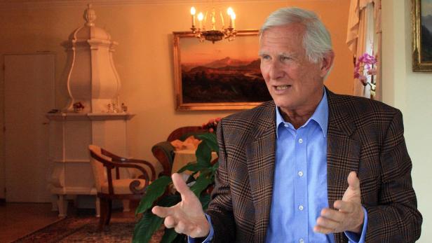 Ein älterer Mann mit blauem Hemd und kariertem Sakko gestikuliert in einem Wohnzimmer.