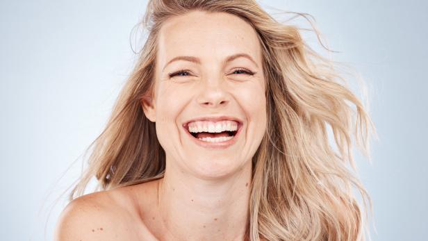 Eine lachende Frau mit blonden, wehenden Haaren vor einem hellblauen Hintergrund.