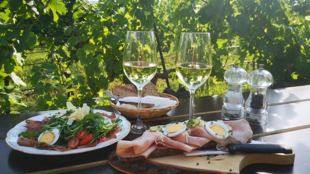 Auf einem Tisch im Weinberg stehen Wein, Brot, Wurst und Salat bereit.