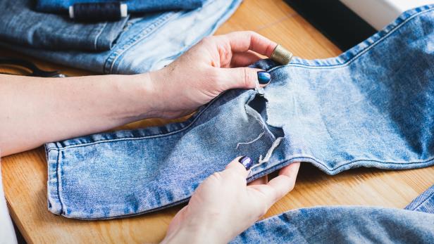 A woman is repairing ripped blue jeans. The concept of economical reuse of things. Homemade needlework.