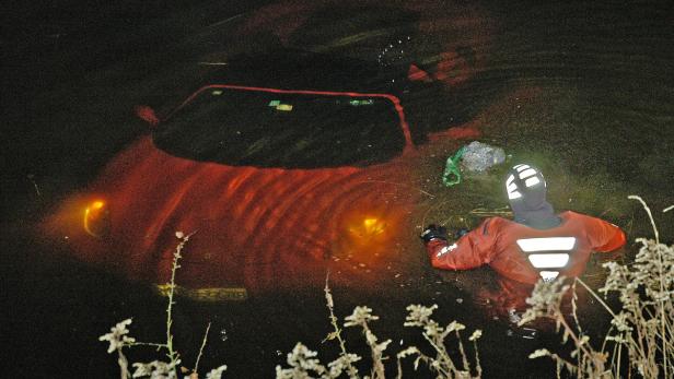 Ein Taucher nähert sich einem roten Auto, das teilweise im dunklen Wasser versunken ist.
