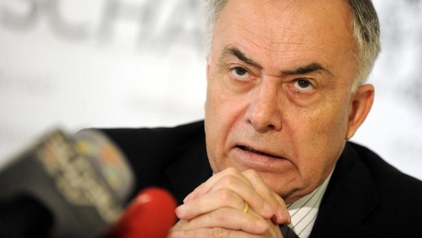 Ein Mann im Anzug mit verschränkten Händen vor Mikrofonen.