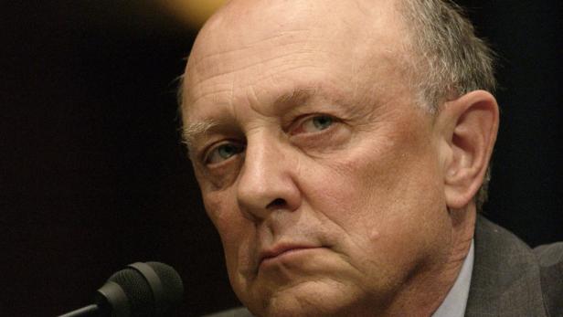 Ein Mann mittleren Alters mit ernstem Gesichtsausdruck steht vor einem Mikrofon.