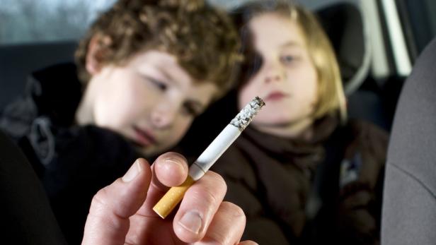 Eine Hand hält eine brennende Zigarette im Auto, im Hintergrund sind zwei Kinder zu sehen.