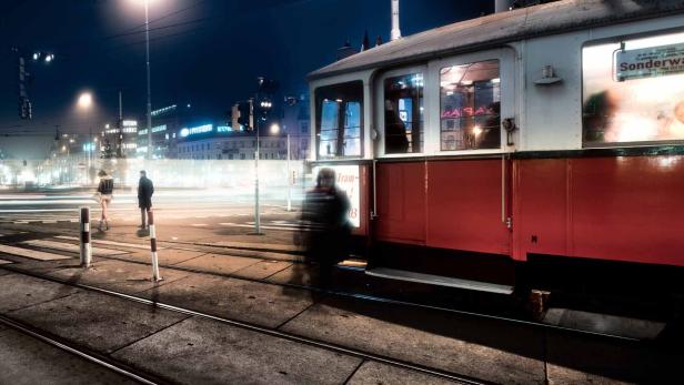 Eine rote und weiße Straßenbahn steht nachts in einer belebten Stadt.