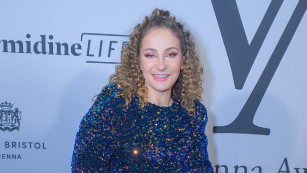 Kristina Vogel mit lockigem Haar sitzt in einem Rollstuhl und trägt ein blaues, glitzerndes Abendkleid vor einer Sponsorenwand.