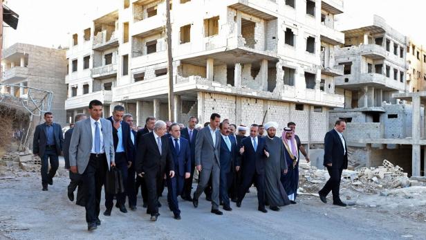 Baschar al-Assad geht mit einer Gruppe von Männern durch eine zerstörte Stadt.