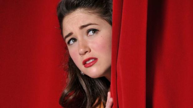 Eine Frau blickt ängstlich hinter einem roten Vorhang hervor.