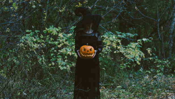 Eine Hexe hält einen geschnitzten Kürbis im Wald.