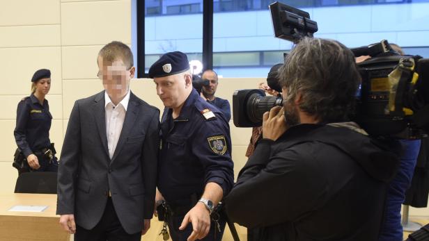 Ein unscharfer Mann in einem Anzug wird von einem Justizbeamten begleitet, während Kameraleute filmen.