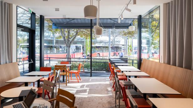 Innenansicht eines Cafés mit Tischen und Stühlen, im Hintergrund fahren Busse vorbei.